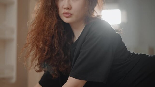 Young woman rests on wooden floor with arm extended and head on forearm, eyes softly open and hair spread across surface, creating peaceful post-exercise calm with warm natural backlight
