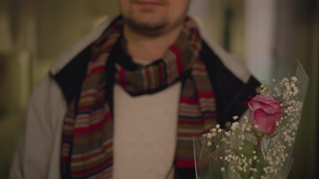 Frontal portrait of man standing in narrow indoor corridor holding wrapped bouquet with single pink rose and baby's breath, facing camera with calm expectant expression, soft warm bokeh glow and