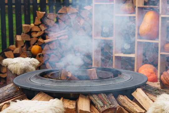 Modern outdoor fire pit, fireplace with smoke surrounded by stacked firewood, soft blankets and autumn decorations. Cozy fall setting for Halloween, Thanksgiving and seasonal outdoor gatherings