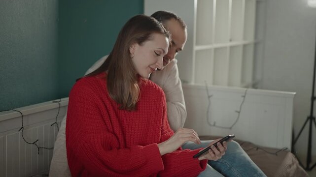 Young woman in red sweater and man sit close on bed, woman extends arm holding smartphone while both lean together and smile toward camera, showing relaxed affectionate home moment