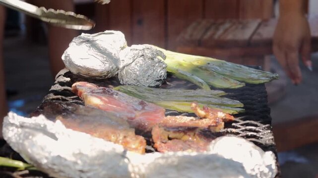 Sizzling carne asada, nopales cactus, and foil-wrapped potatoes cooking on a charcoal grill during a sunny outdoor family barbecue, with tongs turning the meat