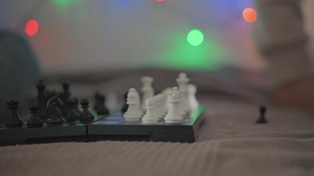 Close view from low angle shows portable chess board during midgame play, with isolated pawn, clustered white and black pieces, blurred festive lights, textured surface, cozy indoor atmosphere, and