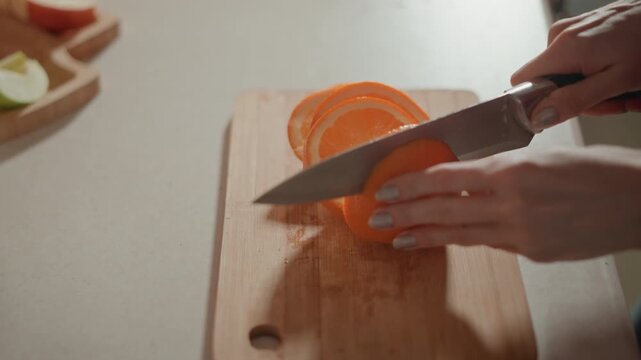 Close-up overhead of hands cutting orange into multiple round slices on wooden cutting board; knife moves downward through fruit leaving layered slices and small puddle of juice on board