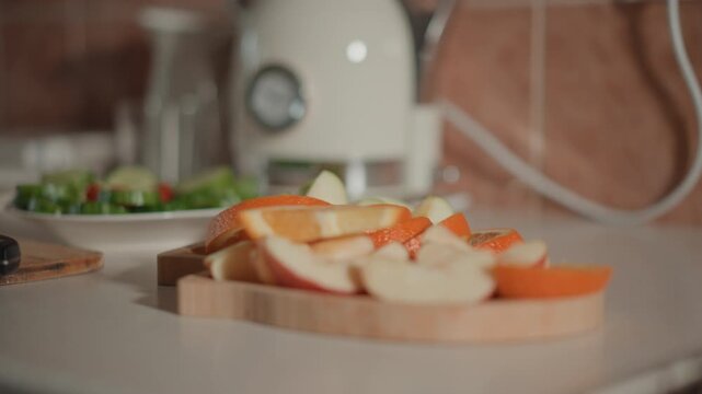 Wooden cutting board holds sliced apples and bright persimmon near plated green salad on pale countertop, with warm kitchen light and shallow focus giving fresh inviting domestic mood