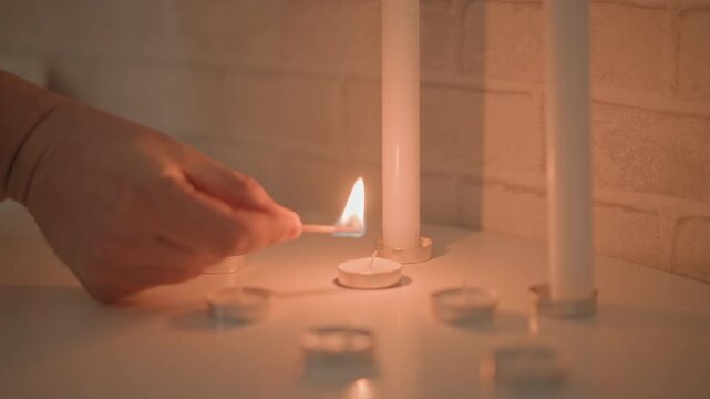 Close view shows hand holding lighter to light tealight wick beside tall white taper candles on glossy table, with warm glow, brick wall backdrop, and soft candle reflections, with hand, lighter