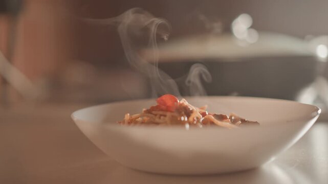 Low-angle view of sauce-coated pasta in shallow white ceramic bowl on neutral kitchen counter, with cherry tomato garnish and visible steam rising for fresh, cozy, appetizing meal styling