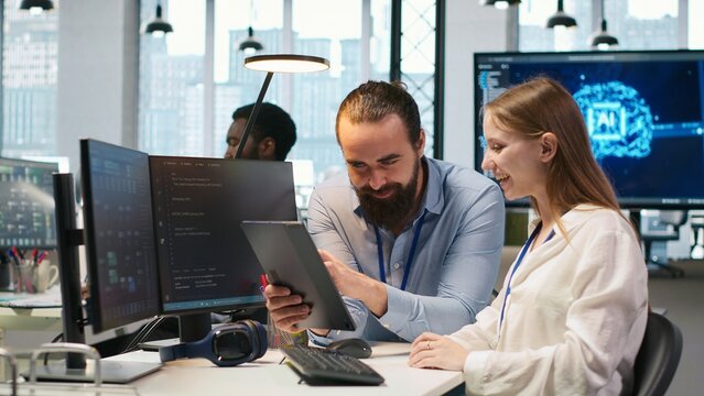 Friendly team leader helps a young engineer code AI algorithms, debug software issues and improve system stability while mentoring the smiling intern at a workstation in a tech startup office.