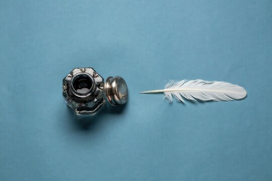 Vintage metal ink bottle with old white quill feather pen on blue background. Antiques for letter writing. Calligraphy tool and historical accessories for creative design.