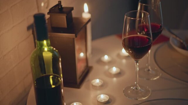 Low angled close view of round dining table featuring two partially filled red wine glasses, upright green wine bottle, lit tealight candles arranged across tabletop, decorative lantern casting warm