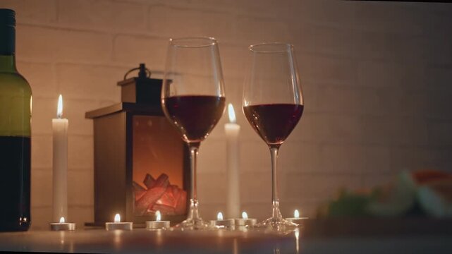 Two filled red wine glasses stand beside upright bottle and decorative lantern on low tabletop, with small candles and string lights casting warm bokeh, soft reflections, intimate cozy evening mood