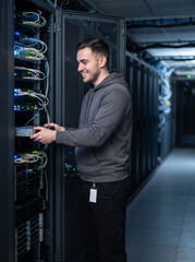 Fototapeta premium Server rack technician in data center expansion with smiling man working on network equipment in dark corridor