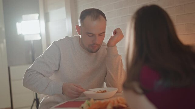 Seated man eats from bowl with focused neutral expression while blurred companion sits across table in warm indoor dining setting, soft window backlight, shallow focus, calm social mood