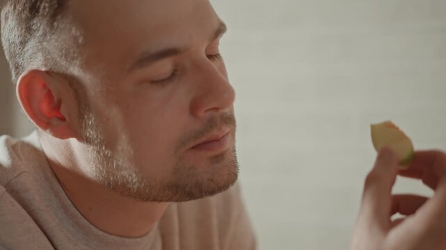 Medium close-up shows adult man in home kitchen lifting bite-sized snack toward lips with focused gaze, relaxed posture, and soft warm light, creating calm everyday food preparation mood