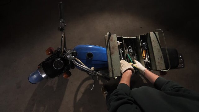 Mechanic working on a classic blue motorcycle, holding an open metal toolbox filled with various hand tools, seen from a top down perspective in a garage setting