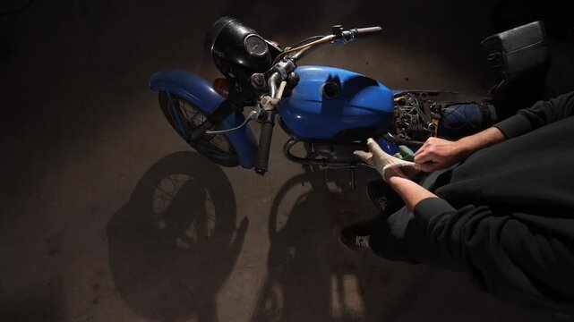 Mechanic with work gloves performing maintenance on a classic blue motorcycle, representing a passion for vintage vehicles, restoration, and skilled craftsmanship in a garage setting
