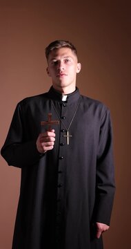 Priest in cassock with wooden cross praying on beige background