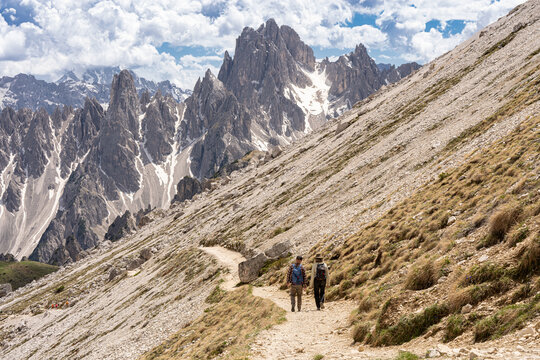 Rear view of two hikers with backpacks walking along a narrow mountain path facing majestic jagged peaks of Cadini di Misurina, high-altitude alpine trekking and active travel concept