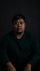 Fototapeta premium A compelling medium shot portrait of a confident Southeast Asian male sitting and looking directly into the camera against a solid dark gray background. The individual is wearing a