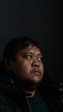 An adult Southeast Asian male with short dark hair looks anxiously upwards while wearing a heavy black winter jacket over a green shirt. The bareheaded Indonesian man features a glistening