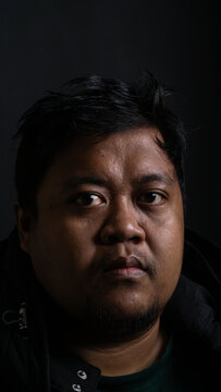 A powerful studio portrait captures an adult Southeast Asian male with dark textured hair staring directly and intensely into the camera lens. The bareheaded Indonesian man wears a