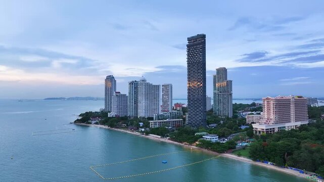 An aerial view of modern high-rise condominiums and resort buildings along the Pattaya coastline with calm blue sea and distant mountains.