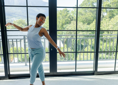 African American woman stretching, dancing in light-blue set at home by glass doors with smartwatch
