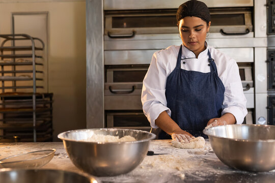 Female baker wearing chef jacket and apron kneading dough in bakery on floured table with bowls