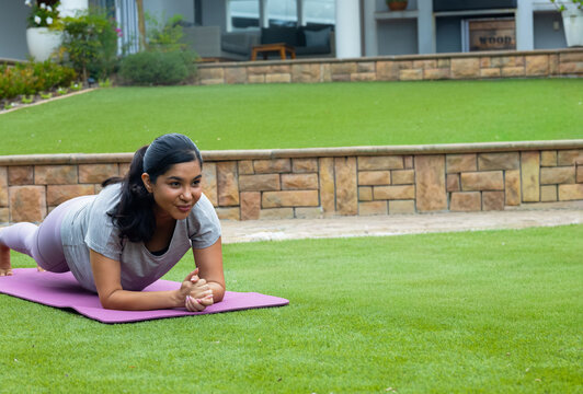 Woman holding forearm plank on purple mat in backyard wearing workout clothes near stone wall