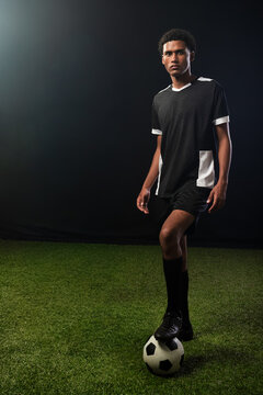 African American male wearing black soccer jersey, cleats standing on studio turf balancing ball