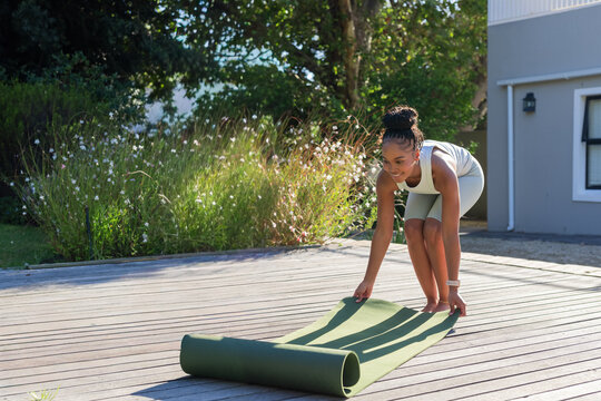 African woman unrolling green yoga mat on backyard wooden deck wearing tank top, copy space