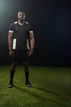 African man standing on green turf in studio wearing black soccer kit and cleats