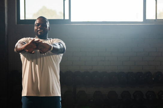 African American man doing forward arm stretch in gym near weights, in light tee and shorts