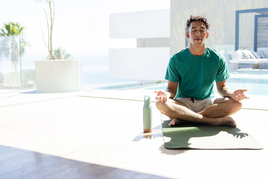 Adult male meditating cross-legged on sunlit terrace by pool with green mat and bottle, copy space