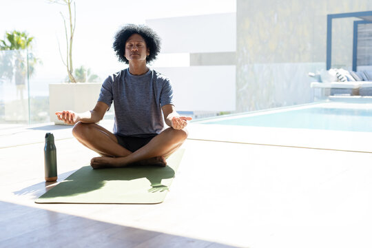 African man sitting meditating cross-legged on green mat by pool in grey tee with water bottle