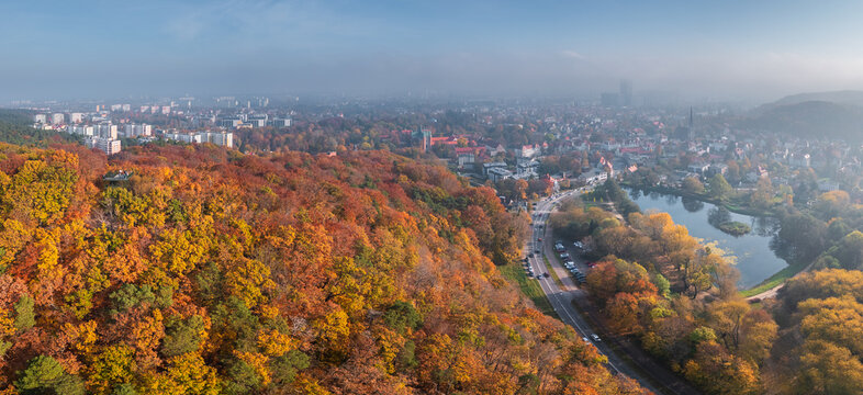 Aerial view of the Oliwa district with vibrant autumn foliage on the hills, a calm pond, and urban buildings under a hazy sky in Gdansk, Pomeranian Voivodeship, Poland.