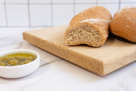 Fresh bread rests on a wooden board. Small bowl holds green olives in olive oil. White tiles frame the clean, bright kitchen. The bread looks soft and freshly baked. Ideal for food, lifestyle