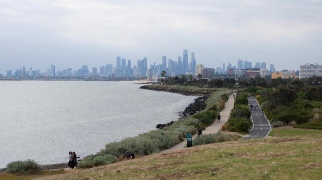 View from Point Ormond Lookout over the Port Phillip Bay coastline, people walking along the coastal path and cycling on the shared bike trail towards the distant Melbourne city skyline in Australia