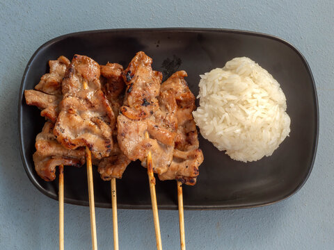 Thai grilled pork skewers (Moo Ping) served with sticky rice on a black plate.