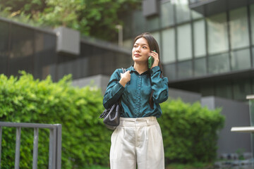 Fototapeta premium Happy young asian businesswoman talking on smartphone standing outdoor modern green office building, Confident freelancer, corporate lifestyle, mobile communication and business network concept