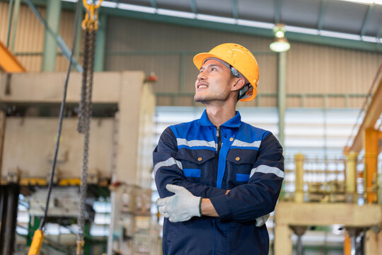 Portrait of confident Asian industrial worker looking up in manufacturing facility, Factory operator standing with arms crossed in industrial plant, Technician looking at overhead equipment in factory