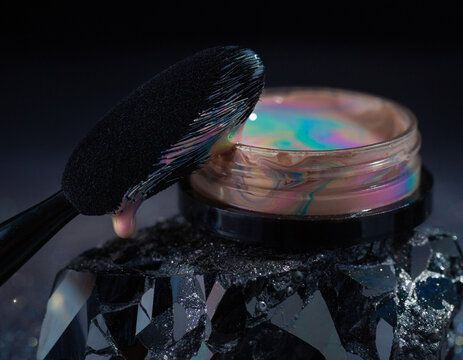 Macro of makeup brush dripping iridescent liquid cosmetic cream on dark crystal.