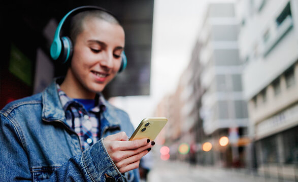 Young woman waiting at bus station while listen music playlist with headphones - Focus on shaved hair girl hand holding mobile phone