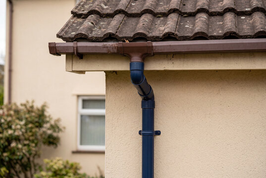 Dark blue plastic downpipe attached to a brown gutter on a house with a tiled roof and beige wall