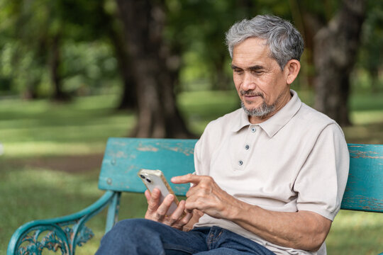 portrait elderly pensioner man spend leisure time relaxing in urban park,sitting on bench,surfing internet on smartphone