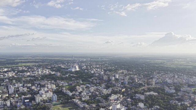 Aerial view of Kushtia city featuring a mix of small and large buildings, residential neighborhoods, extensive tree cover, and an open blue sky with white clouds.