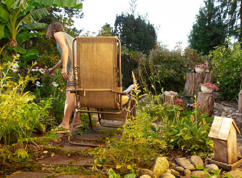 Woman enjoying peaceful moment tending to lush plants in vibrant backyard garden with cozy brown lounger chair, wooden birdhouse, potted flowers on tree stumps, stone path, tropical banana palm trees,