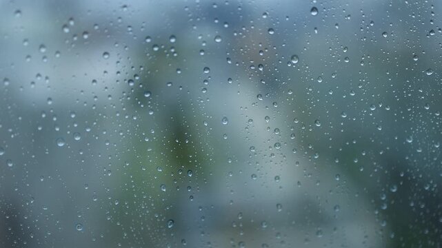 A close-up of rain droplets and water streaks on a window glass with a blurred green exterior visible outside on a rainy day.