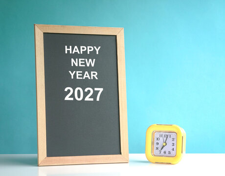 Happy New Year 2027 message on a chalkboard next to yellow clock.