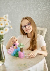 Fotobehang Lego Smiling girl wearing glasses plays with colorful magnetic building blocks at a table at home, enjoying a creative construction activity that develops imagination, learning and problem-solving skills.  © Yulia Mihaylova