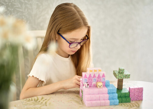 Focused girl wearing glasses is playing with colorful magnetic blocks, building a pink castle and tree at home during an educational construction activity at a table in a bright, cozy interior.